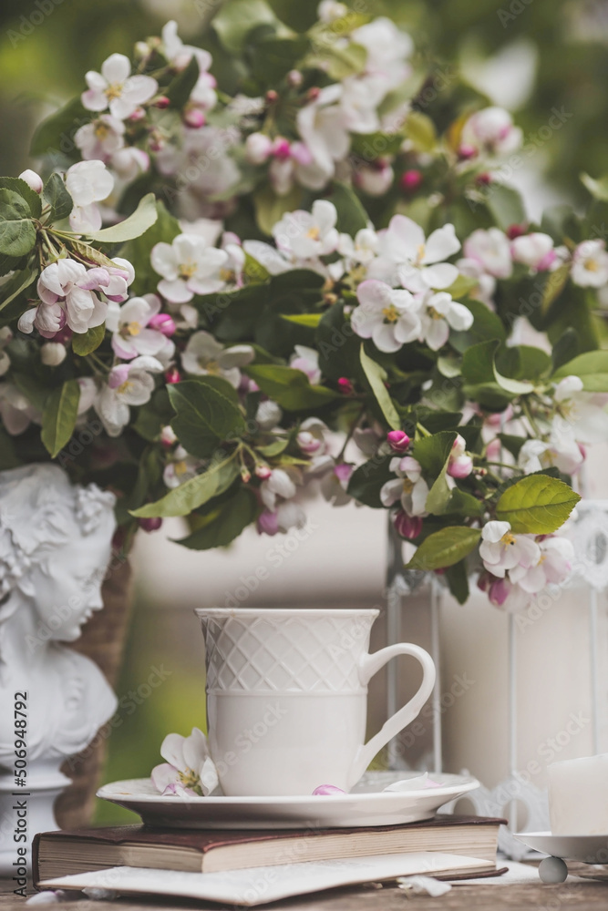 A beautiful postcard. A white coffee cup with a saucer, a statuette, candles, a book and a vase with a bouquet of blooming apple trees. Beautiful still life. Spring time. The concept of 