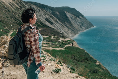 an adult woman with a backpack and in a plaid shirt went hiking in the mountains near the ocean on a spring sunny day, a pensioner admires the sea in good weather