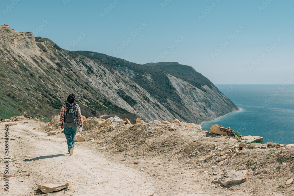 an adult woman with a backpack and in a plaid shirt went hiking in the mountains near the ocean on a spring sunny day, walks along the road with a bottle in her hands. View from the back
