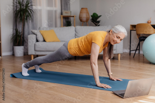 Happy sporty senior woman standing in plank on yoga mat, working out to online sports video at home