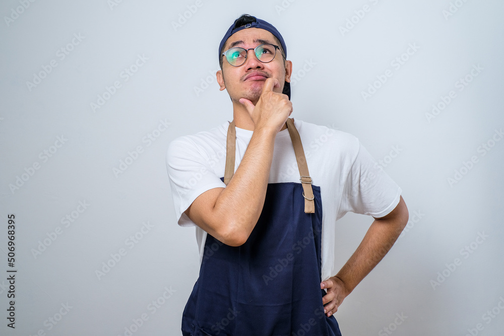 Portrait of attractive Asian barista man in blue apron thinking about question with hand on chin Portrait of attractive Asian barista man in blue apron thinking about question with hand on chin