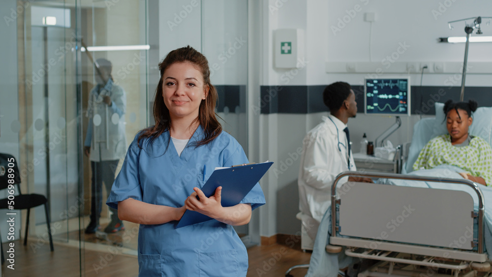 Obraz premium Portrait of woman working as nurse holding checkup papers while standing in hospital ward. Medical assistant with documents looking at camera and smiling, giving help to specialist.