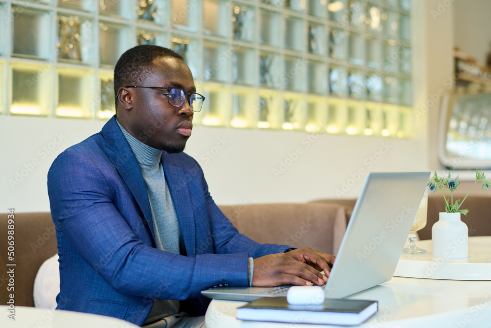African freelancer in eyeglasses sitting at table and typing on laptop, he working online at cafe African freelancer in eyeglasses sitting at table and typing on laptop, he working online at cafe
