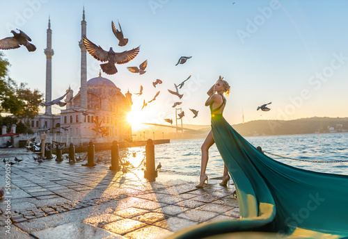 Woman in long green dress posing with flying pigeons at the historical square near Ortaköy mosque and Bosporus waters