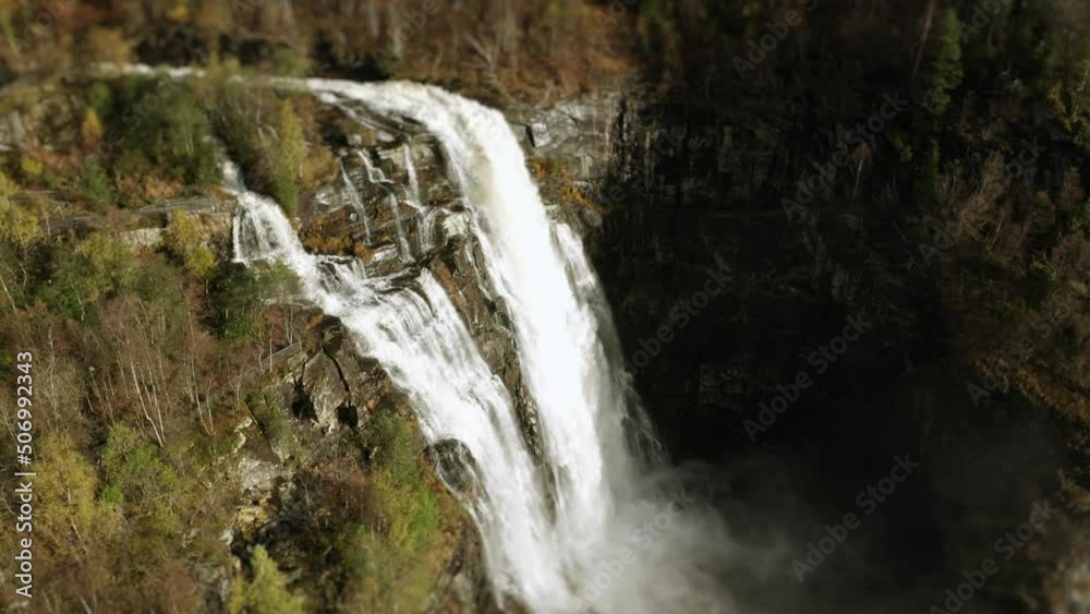 Aerial view of the stunning Skjerfossen waterfall. A powerful torrent of whitewater falls from the cliffs. Water spray rising high in the air. A narrow road below. Tilt-shift. Pan right. Aerial view of the stunning Skjerfossen waterfall. A powerful torrent of whitewater falls from the cliffs. Water spray rising high in the air. A narrow road below. Tilt-shift. Pan right.