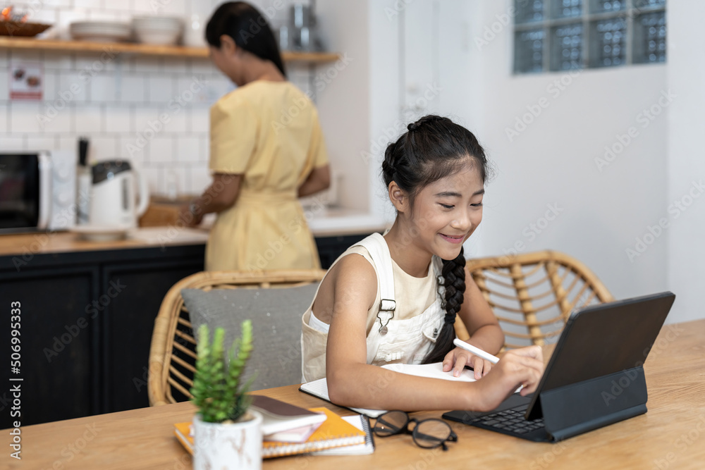 Portrait of little girl using laptop during online class with tutor or teacher while sitting at desk in cozy kitchen interior with mother in background Portrait of little girl using laptop during online class with tutor or teacher while sitting at desk in cozy kitchen interior with mother in background