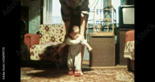 Mother helping child to walk in room. First baby steps, mother holding child in Soviet apartment interior. Vintage color film. Cinema lens flare. Retro 80s, 90s. Family home archive 8mm. 1980s-1990s photo