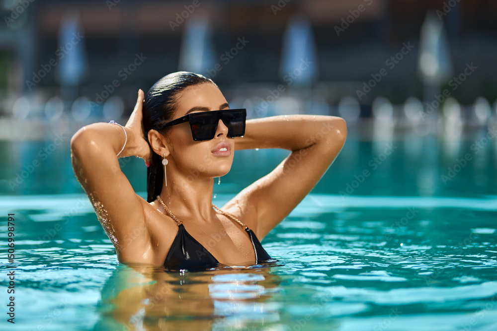 Gorgeous brunette girl in sunglasses and swimsuit relaxing in turquoise pool water. Portrait view of beautiful lady holding hands over head while having fun in pool of luxury hotel. Concept of relax.