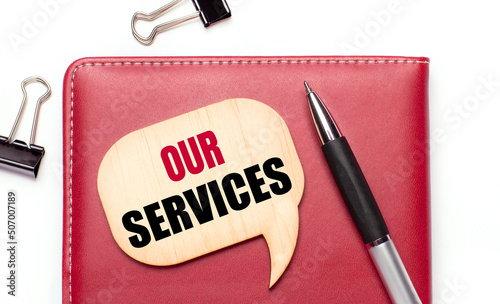 On a light background there are black paper clips, a pen, a burgundy notepad a wooden board with the text OUR SERVICES