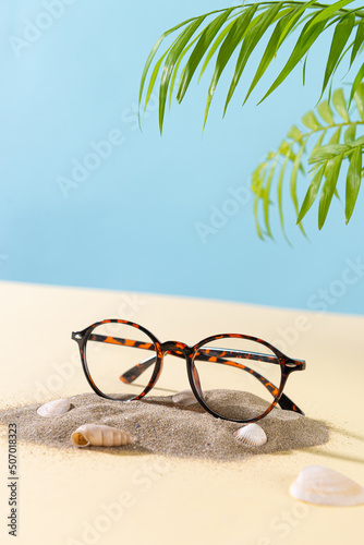 Trendy Eyeglass frame on beach with palm leaves, trendy Still Life Style. Tortoiseshell frame glasses on sand. Optic store advertisement background. Vertical. Close up. Glasses sale concept. Eyeweare