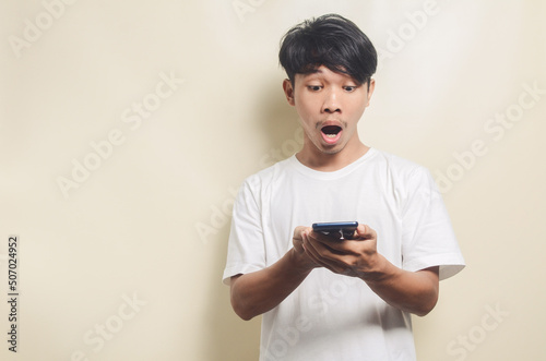 asian man wearing white t-shirt with surprised gesture after getting a message by phone on isolated background