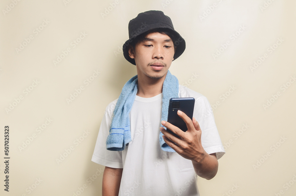 asian man wearing bright white t-shirt in black hat with blue towel carrying phone on isolated background asian man wearing bright white t-shirt in black hat with blue towel carrying phone on isolated background