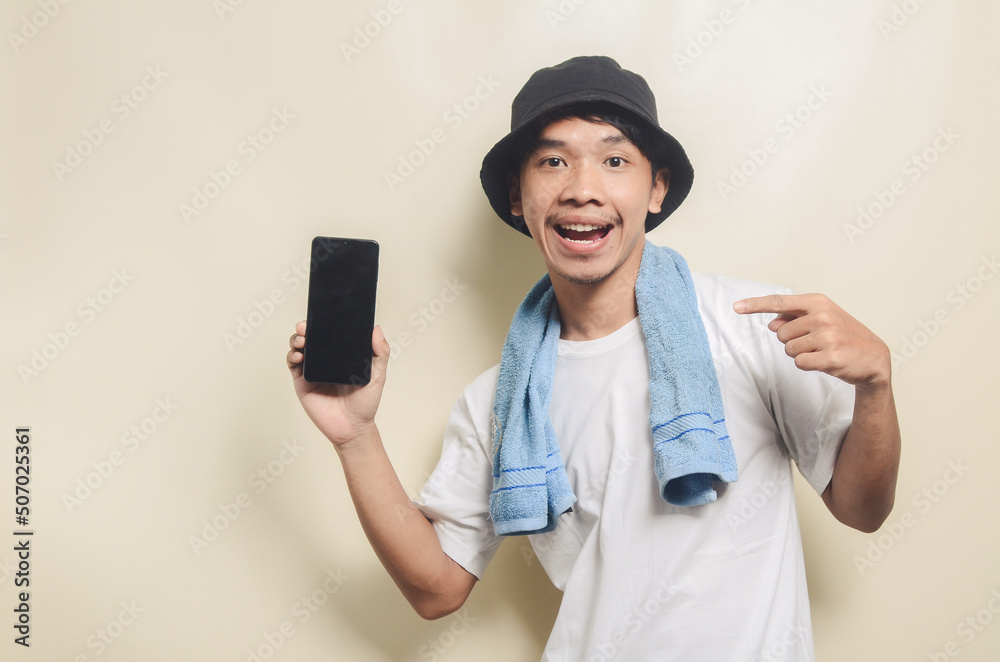 asian man wearing bright white t-shirt in black hat with blue towel pointing at phone screen on isolated background asian man wearing bright white t-shirt in black hat with blue towel pointing at phone screen on isolated background