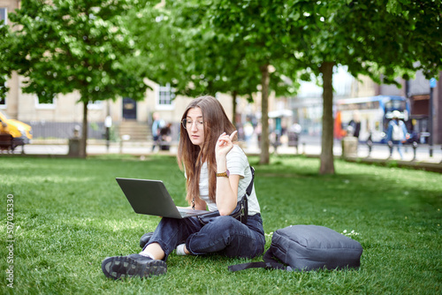A young pretty woman sits on the lawn with a laptop. A woman works remotely with a laptop outdoors in the park. The concept of remote work. Freelance work.