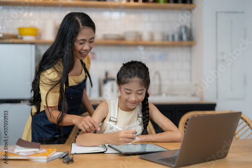 Asian mother and little daughter sit at desk in kitchen studying online together, biracial mom and small girl child handwrite, do homework, learning at home, homeschooling concept © Natee Meepian