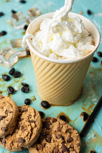 Aerial view of a cardboard cup with coffee and cream on a blue table with spilled coffee, ice, cookies and coffee beans, vertical
