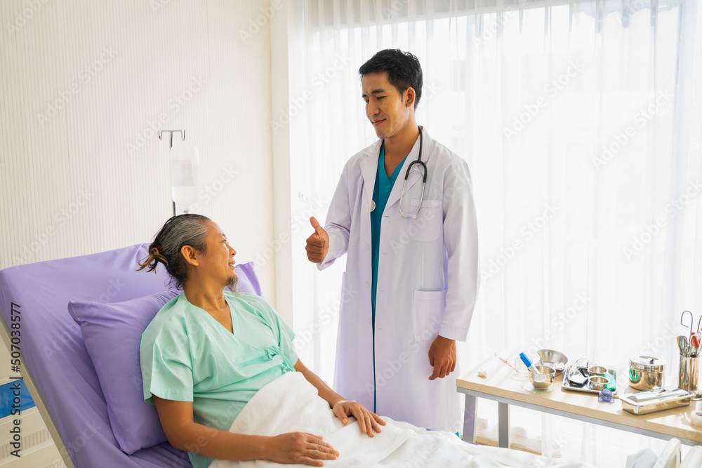 Doctor with stethoscope shows thumb up to elderly patient man with saline sitting on bed. Doctor or nurse take care of senior patient health admission at hospital Doctor with stethoscope shows thumb up to elderly patient man with saline sitting on bed. Doctor or nurse take care of senior patient health admission at hospital