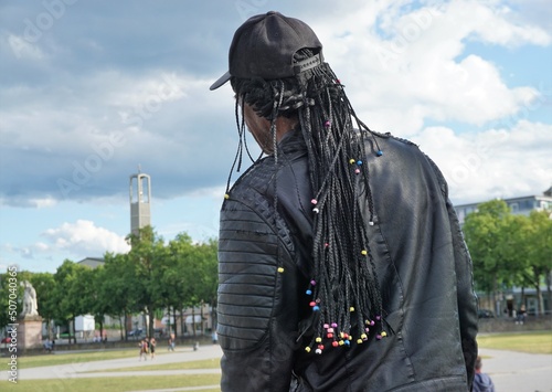 Hinterkopf und Rücken von schwarzhäutigem Mann mit schwarzer Kappe, langen schwarzen Rastafari-Locken über schwarzer Lederjacke vor Platz mit grüner Wiese, Bäumen, Gebäuden, Himmel und Wolken  photo