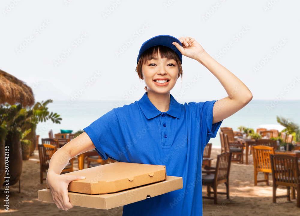 food, service and job concept - happy smiling delivery woman in blue uniform with takeaway pizza boxes over open air restaurant on beach background