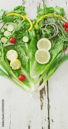 Vertical image of fresh tomatoes and green vegetables forming heart on white rustic background