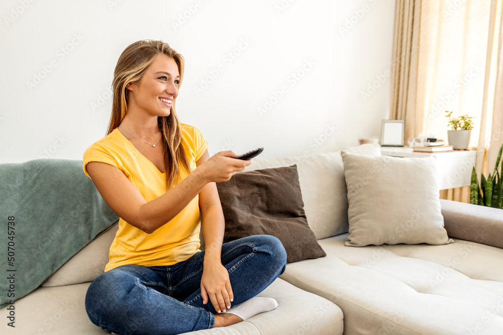 Shot of a young woman holding a remote control while sitting on the sofa at home. Young woman spending a relaxing weekend at home watching tv. Woman watching TV while relaxing on her sofa at home. Shot of a young woman holding a remote control while sitting on the sofa at home. Young woman spending a relaxing weekend at home watching tv. Woman watching TV while relaxing on her sofa at home.