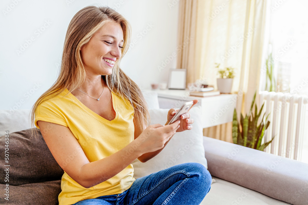 Attractive young woman sitting on her couch and using her cellphone while at home. Smiling young woman using mobile phone app playing game, shopping online, ordering delivery relax on sofa. Attractive young woman sitting on her couch and using her cellphone while at home. Smiling young woman using mobile phone app playing game, shopping online, ordering delivery relax on sofa.