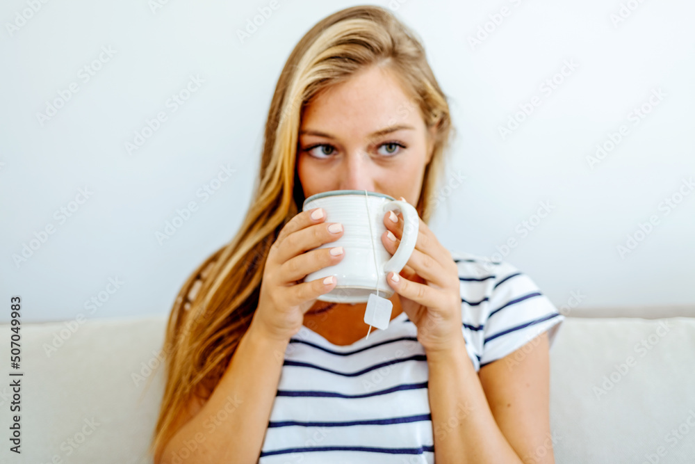 Thoughtful woman sitting on couch holding a cup of tea in living room. Young woman drinking a cup of tea while relaxing on sofa at home. Portrait of girl with hot drink in hands, looking away. Thoughtful woman sitting on couch holding a cup of tea in living room. Young woman drinking a cup of tea while relaxing on sofa at home. Portrait of girl with hot drink in hands, looking away.