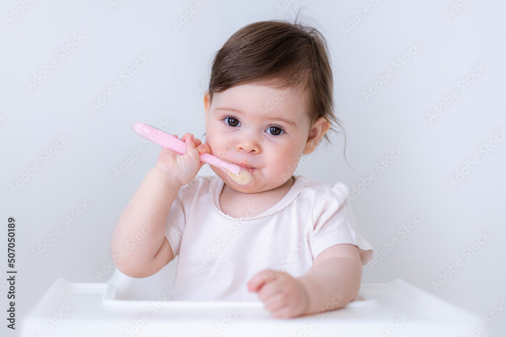 Cute Baby girl eating blend mashed food sitting, on high chair, mother feeding child, hand with spoon for vegetable lunch, baby weaning, first solid food for young kid. Cute Baby girl eating blend mashed food sitting, on high chair, mother feeding child, hand with spoon for vegetable lunch, baby weaning, first solid food for young kid.