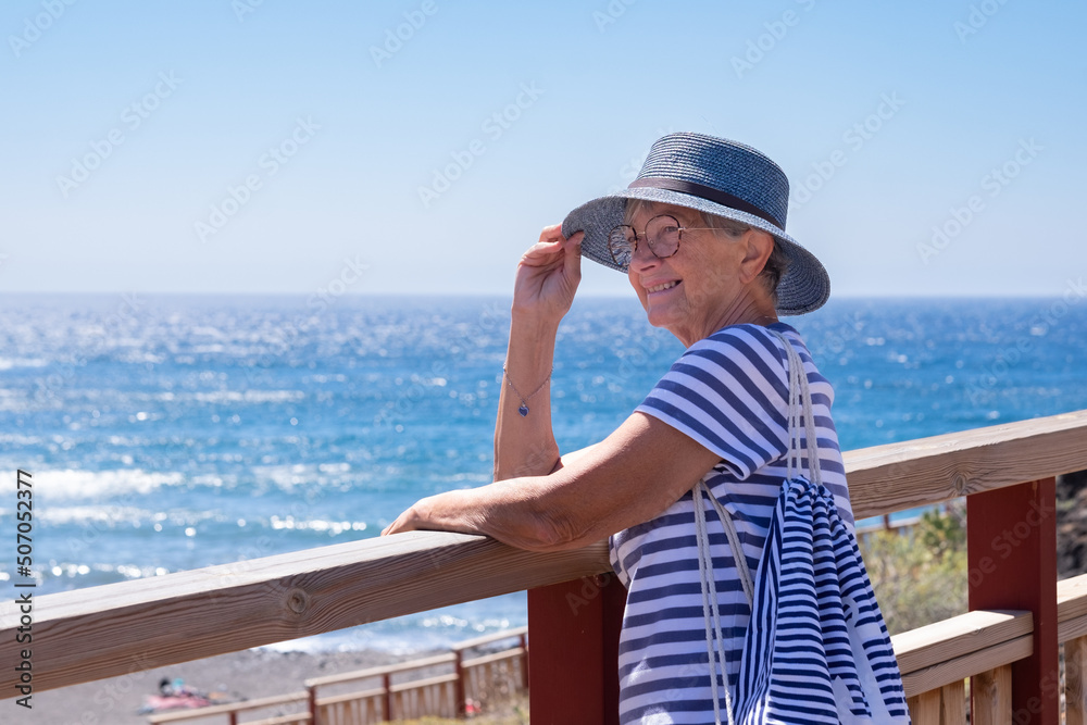 Happy senior woman dressed in blue walking along the sea holding her hat lest it fly away. Mature lady with eyeglasses enjoying freedom and relax in vacation