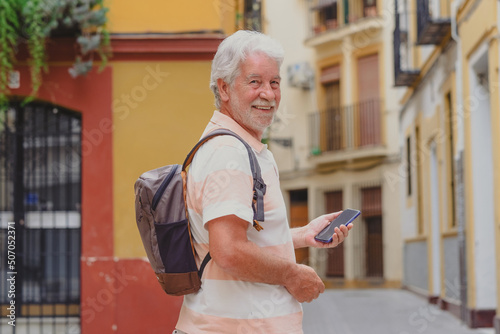 Happy senior tourist man holding backpack on shoulder walking in the alleys of the old town in Seville, Spain looking at camera with phone in hand