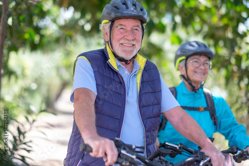 Portrait of happy active caucasian senior couple with electric bicycles running outdoors in the park. Two smiling seniors wearing cycling helmet enjoying a healthy lifestyle in nature #507052388