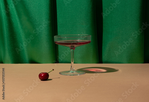 Champagne glass, cherry, rich green drapery. Creative fruity cocktail arrangement. 60s vibes. 