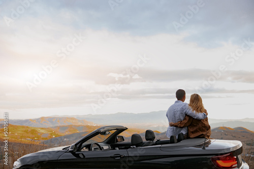 Happy couple man and woman hugging enjoying beautiful mountains standing near cabriolet, back view