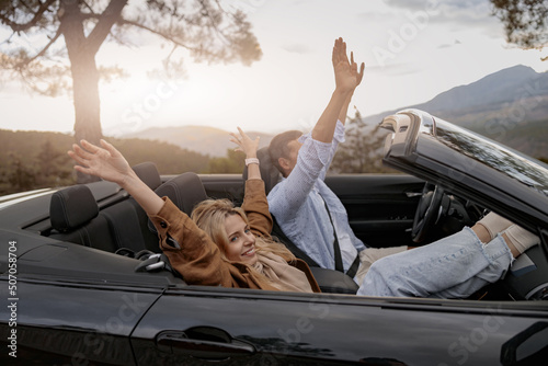 Side view of cheerful boyfriend and girlfriend sit in convertible car in mountains. Vacation concept