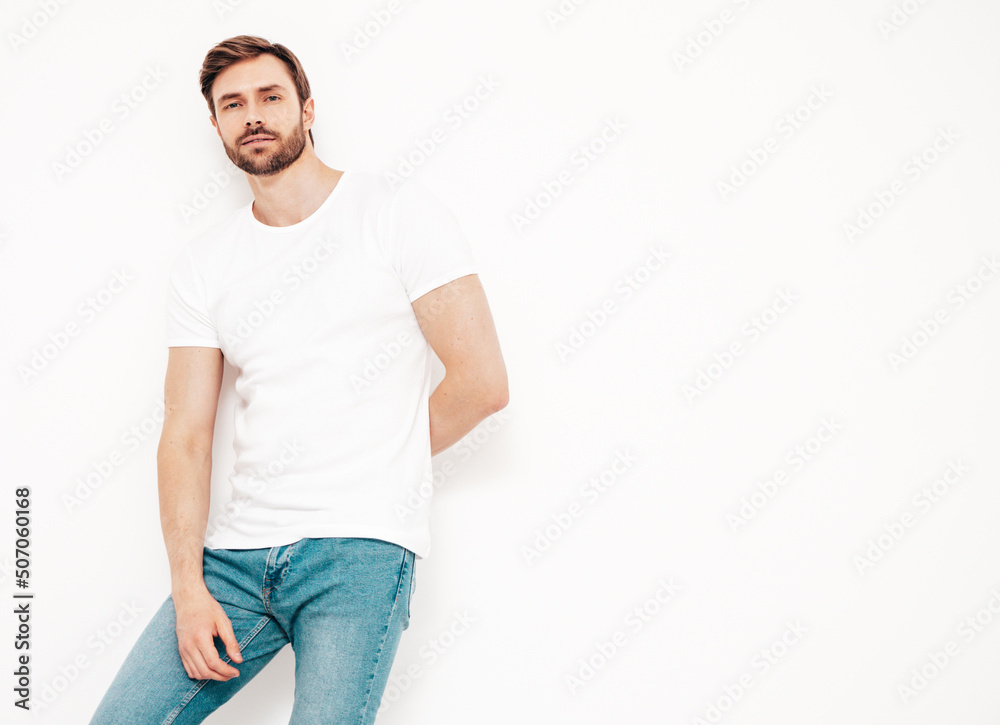 Fototapeta premium Portrait of handsome smiling stylish hipster lambersexual model. Sexy man dressed in T-shirt and jeans. Fashion male isolated on white wall in studio