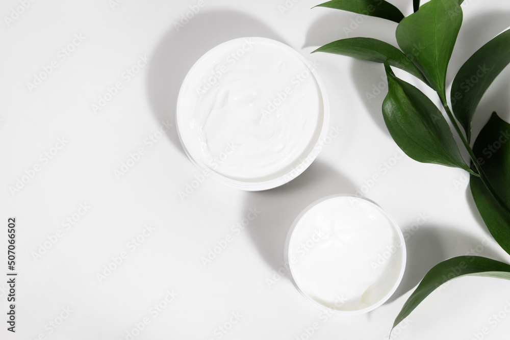 A mock up white jar of cream on a white gray background, on a white table with green leaves of a tropical plant, with hard shadows. Stylish look of the product, identity.
