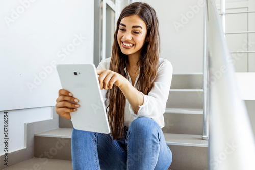 Young beautiful Caucasian female college student using tablet on a staircase. Improving online visibility with modern technology. Happy young woman using digital tablet