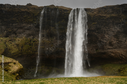 Shiny white waterfall frontal in center from bottom over dark cliff  foreground green-yellow meadow  sky monotonous grey