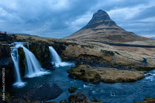 Icelanding tourist attraction with three waterfalls left side, pebble stones and dark calm river in front, and impressive shark fin shaped mountain in background, yellow autumn colours, cloudy bluish © F6 Photography