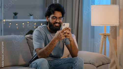 Smiling Arabian Indian man bearded male looking in smartphone smile texting with friend girlfriend messaging chatting at home sitting on couch at night playing game in mobile app watching funny video