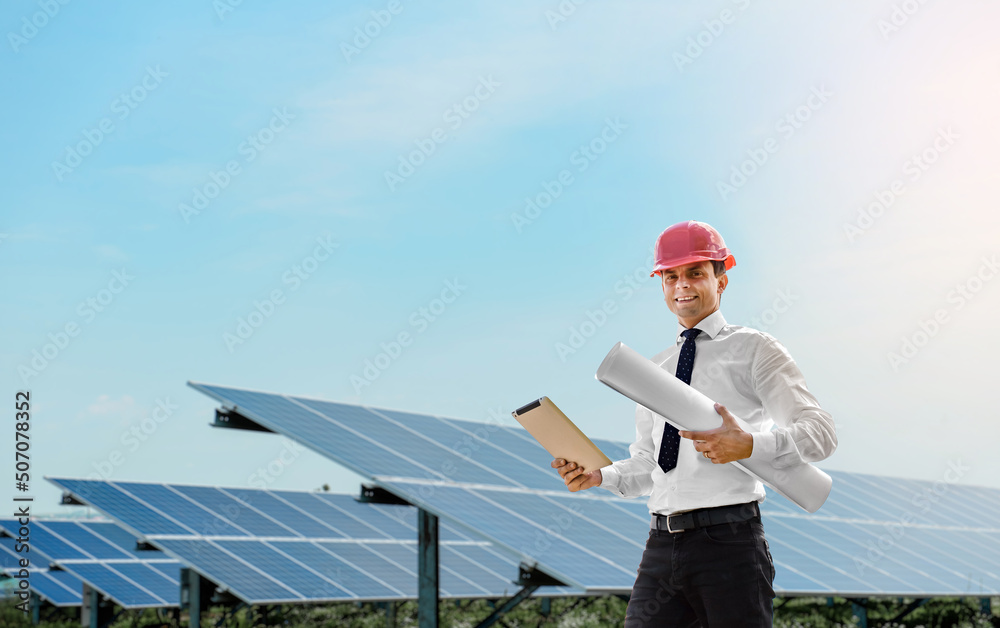 Young man engineer in white shirt and red safety helmet holding a tablet and project paper plan and  verifying the photovoltaic panels at the solar power plant.