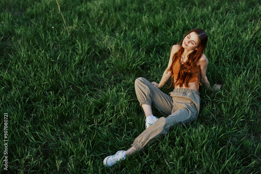 Fototapeta premium A girl sits on the grass in the park and enjoys the beautiful view of the summer sunset