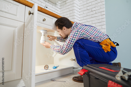 Фотография Skilled male worker plumbing repair service installs siphon under sink in kitchen in apartment