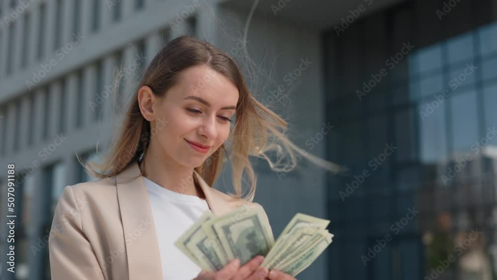 Happy caucasian female worker counting cash on street with office building on background. Smiling businesswoman holding dollars banknotes in hands and enjoying company income. Happy caucasian female worker counting cash on street with office building on background. Smiling businesswoman holding dollars banknotes in hands and enjoying company income.