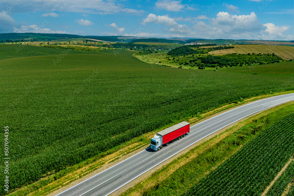 red truck driving on asphalt road on the highway. road through a beautiful green field on a background of blue clouds. seen from the air. Aerial view landscape. drone photography. cargo delivery