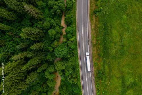 white truck driving on asphalt road on the highway. road through beautiful green forest. seen from the air. Aerial top view landscape. drone photography. cargo delivery