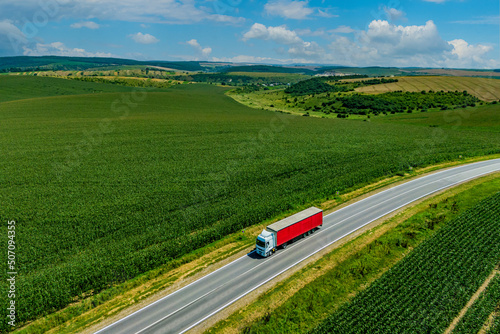 red truck driving on asphalt road on the highway. road through a beautiful green field on a background of blue clouds. seen from the air. Aerial view landscape. drone photography. cargo delivery