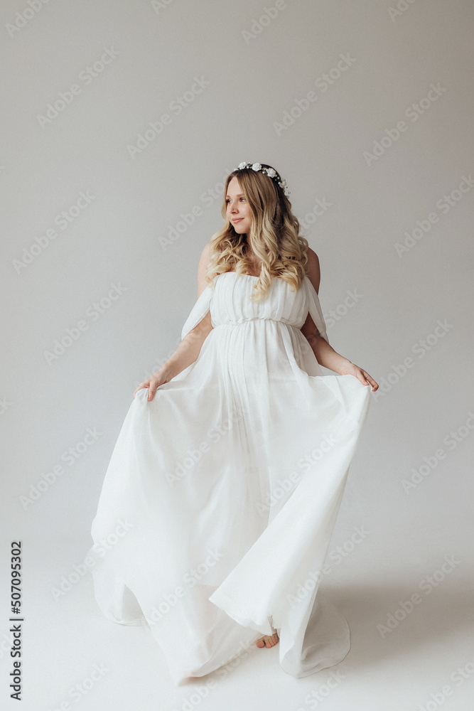 Blonde pregnant woman in a white dress with a wreath on her head in the studio on a white background holds her hand on her stomach Blonde pregnant woman in a white dress with a wreath on her head in the studio on a white background holds her hand on her stomach