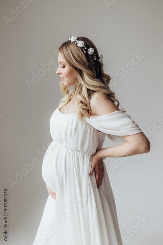 Blonde pregnant woman in a white dress with a wreath on her head in the studio on a white background holds her hand on her stomach