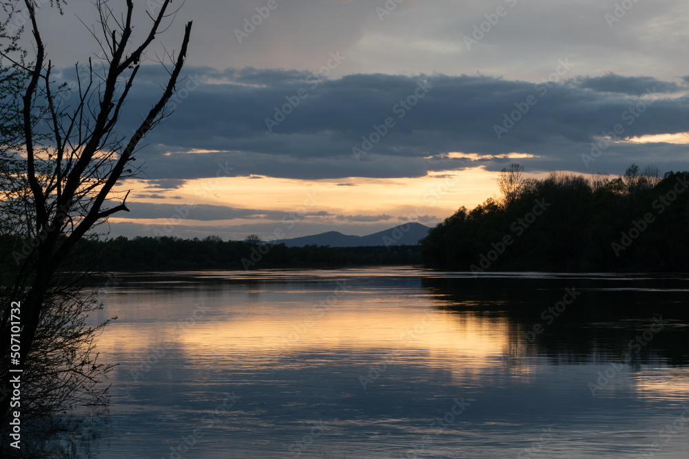 Landscape of river and mountain silhouette with cloudy sky, Sava river with forested shore and Motajica mountain during sunset with glow in clouds